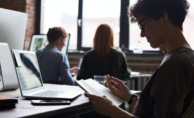 Woman at work writing in note book, slightly in silhouette with other people working behind